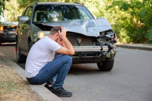 A man sitting on the side of the road in front of his smashed car, wondering if he needs a lawyer after a head-on collision in New Orleans.
